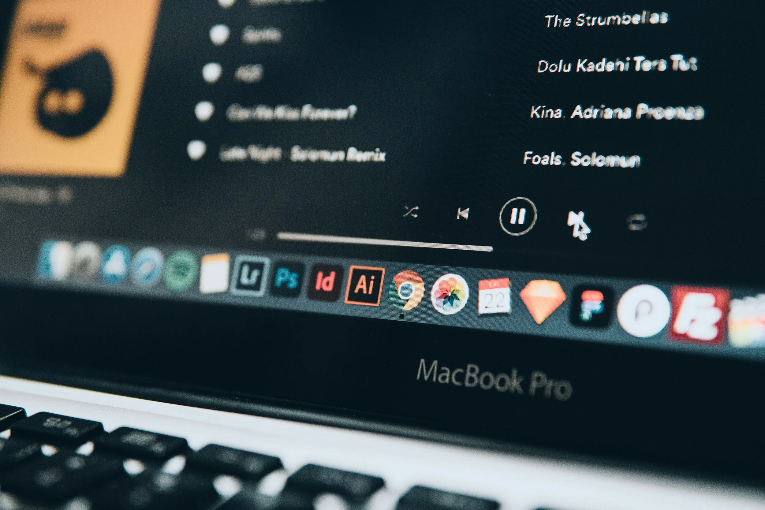 Close-up of a MacBook Pro screen displaying a music playlist and app icons on the dock, including Chrome, Spotify, Adobe Illustrator, and others.