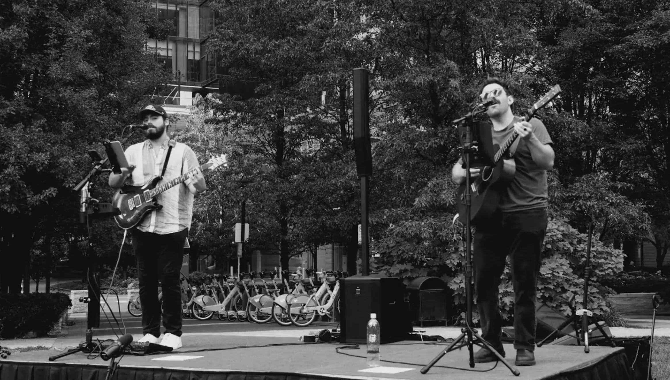 Two male musicians performing on an outdoor stage; one is playing an electric guitar, the other a classical guitar.