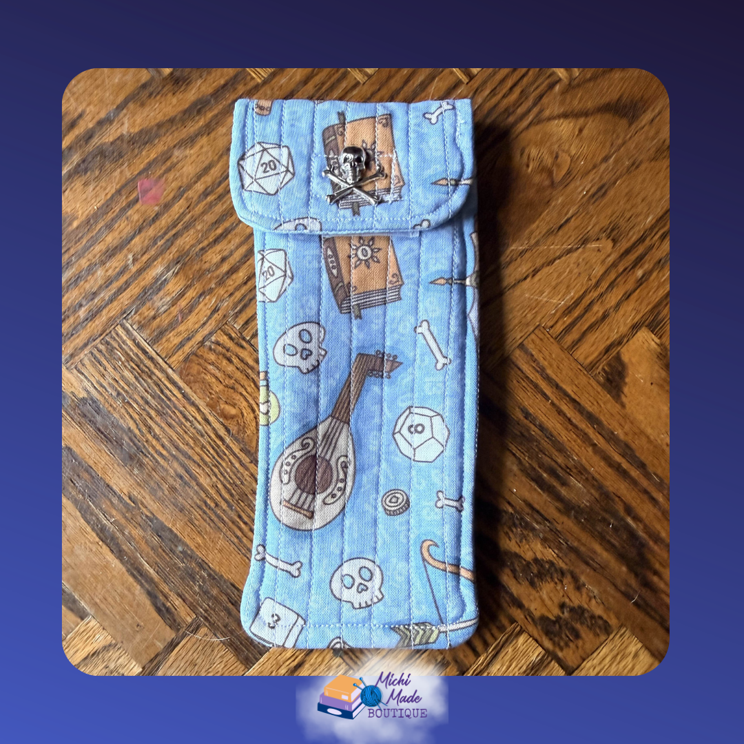 Choose Your Class Roll D20. Quilted eyeglass case in sky blue with TTRPG icons incl. D20 dice, scrolls, a lute, skulls, and bones, fastened by a silver skull and crossbones decorative button.