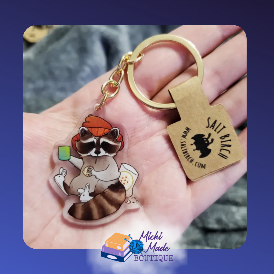 Cozy raccoon keychain in beanie, holding drink with snacks.