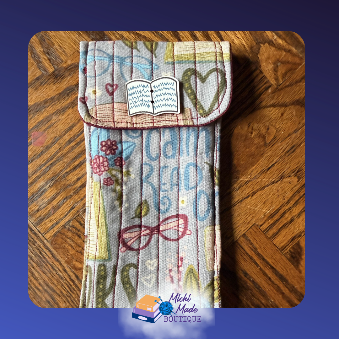 Open Book Reading Life. Quilted eyeglass case in soft multicolor book fabric with reading glasses,  flowers, & books in blue, pink, and yellow, with a white open book decorative button.