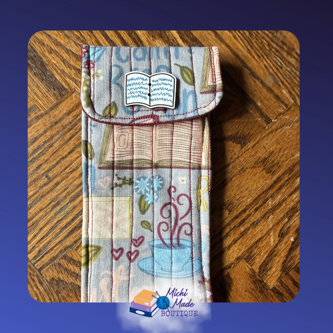 Open Book Tea & Pages. Quilted eyeglass case in soft multicolor bookish fabric with stacked books, a teacup, blue florals, and heart swirls, with a white open book decorative button.