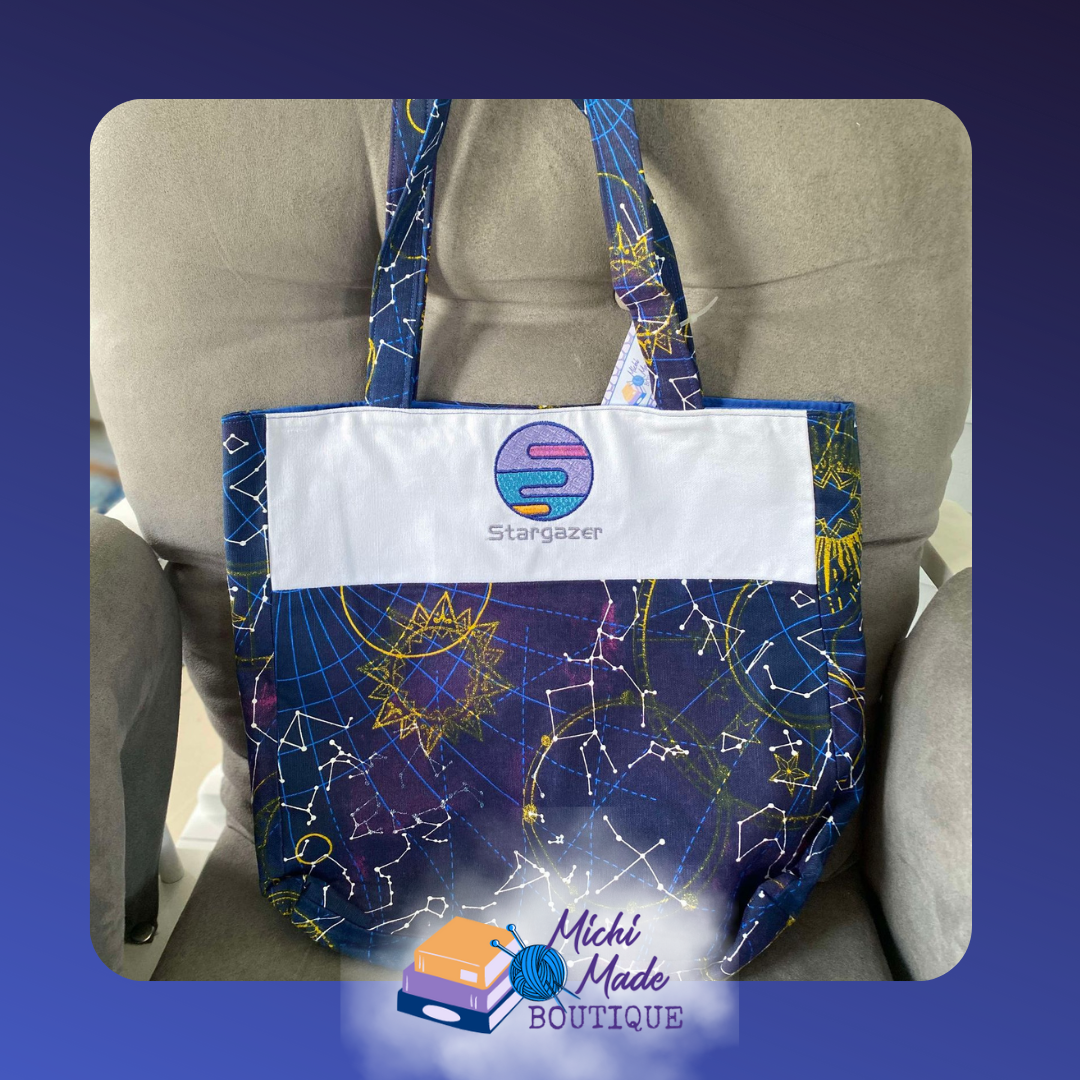 Stargazer Embroidered Tote Bag.png Deep navy constellation print tote bag with a white fabric panel featuring a planet emblem embroidered in pink, teal, and blue with the word Stargazer below.