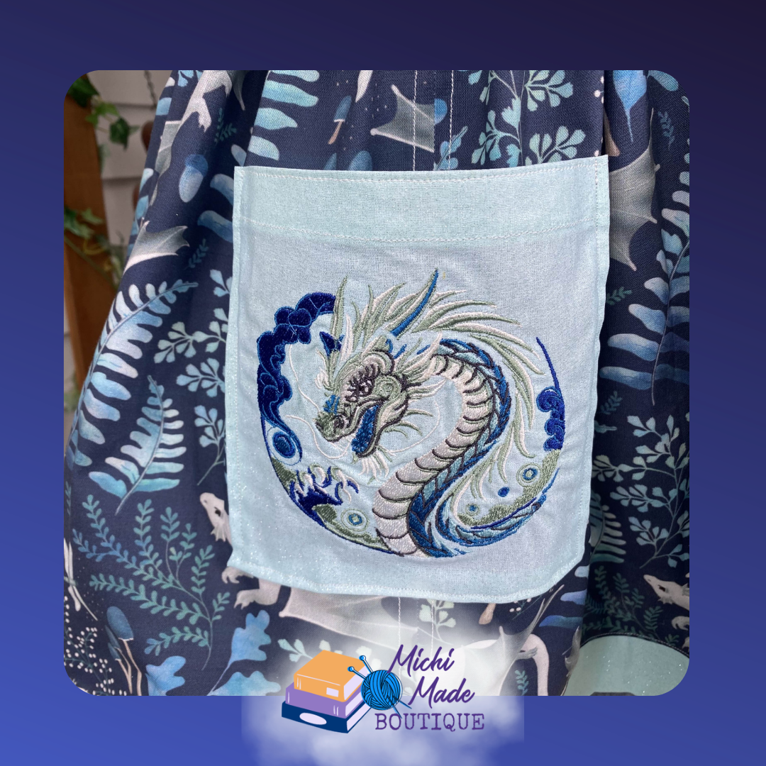 Close-up of a light blue apron pocket with a circular embroidery of a coiled Eastern-style dragon in ivory, navy, sage, and teal thread, surrounded by swirling clouds and decorative scroll details.