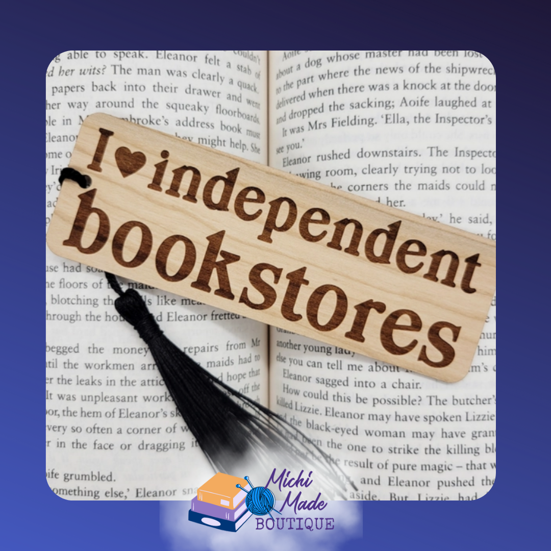 Wooden bookmark engraved with “I ♥ independent bookstores,” black tassel, on open book.