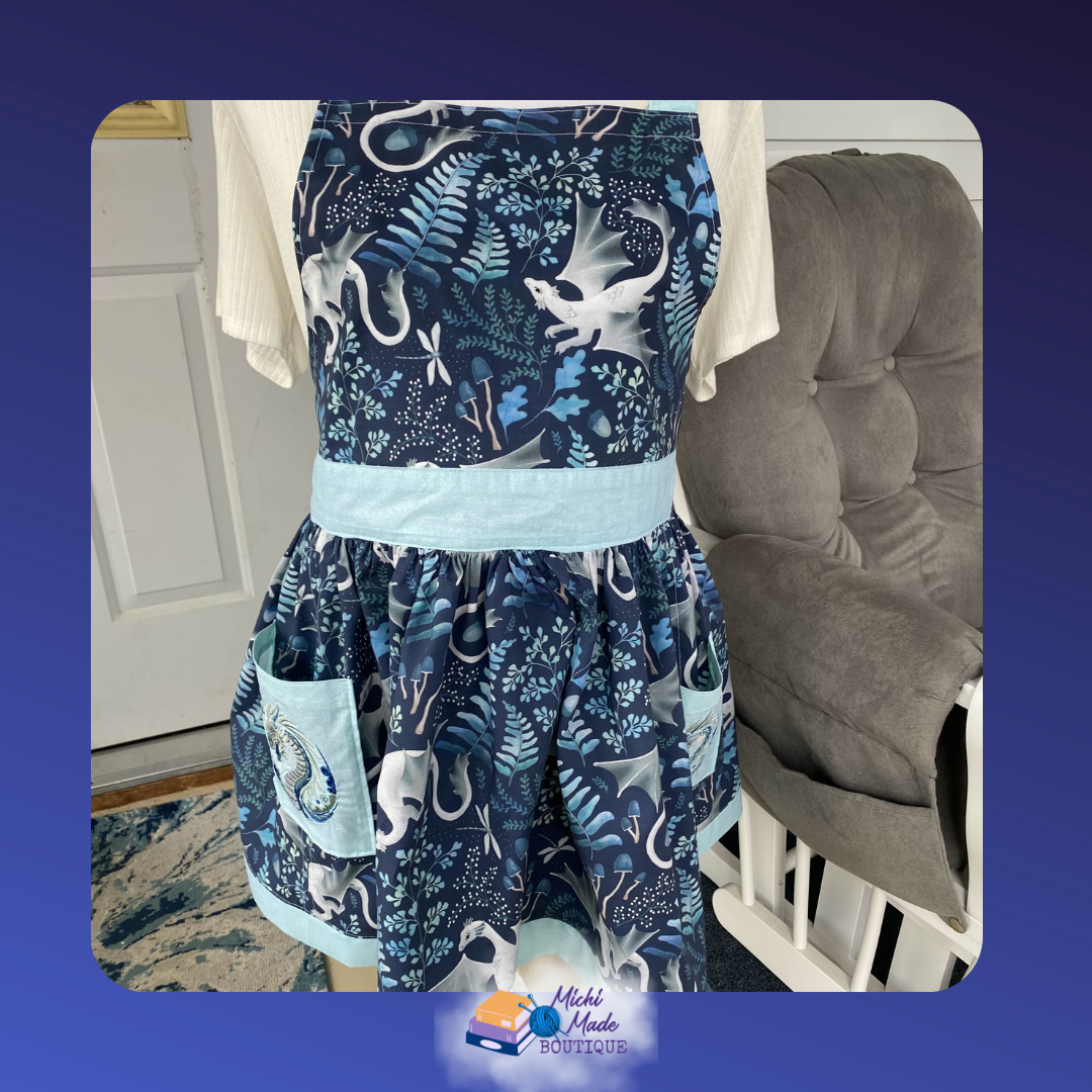 Navy blue apron with white and teal dragons among botanical foliage, light blue waistband, gathered skirt, and two embroidered light blue pockets.