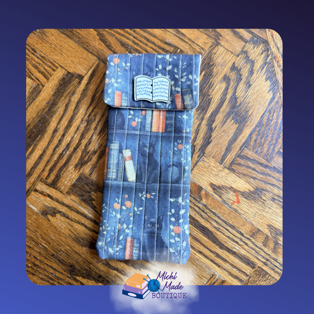 Cats and Books Eyeglass Case.png Quilted eyeglass case in dark teal with an illustrated bookshelf print of colorful books and orange florals, decorated with a white open book button.