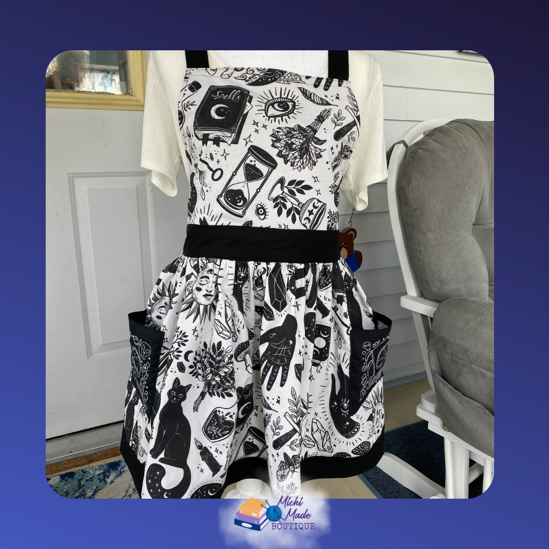 Witchy Black Cat Apron.png White apron with black witchcraft print including a black cat, spell book, crystals, and hourglass. Black waistband, gathered skirt, and two black pockets.