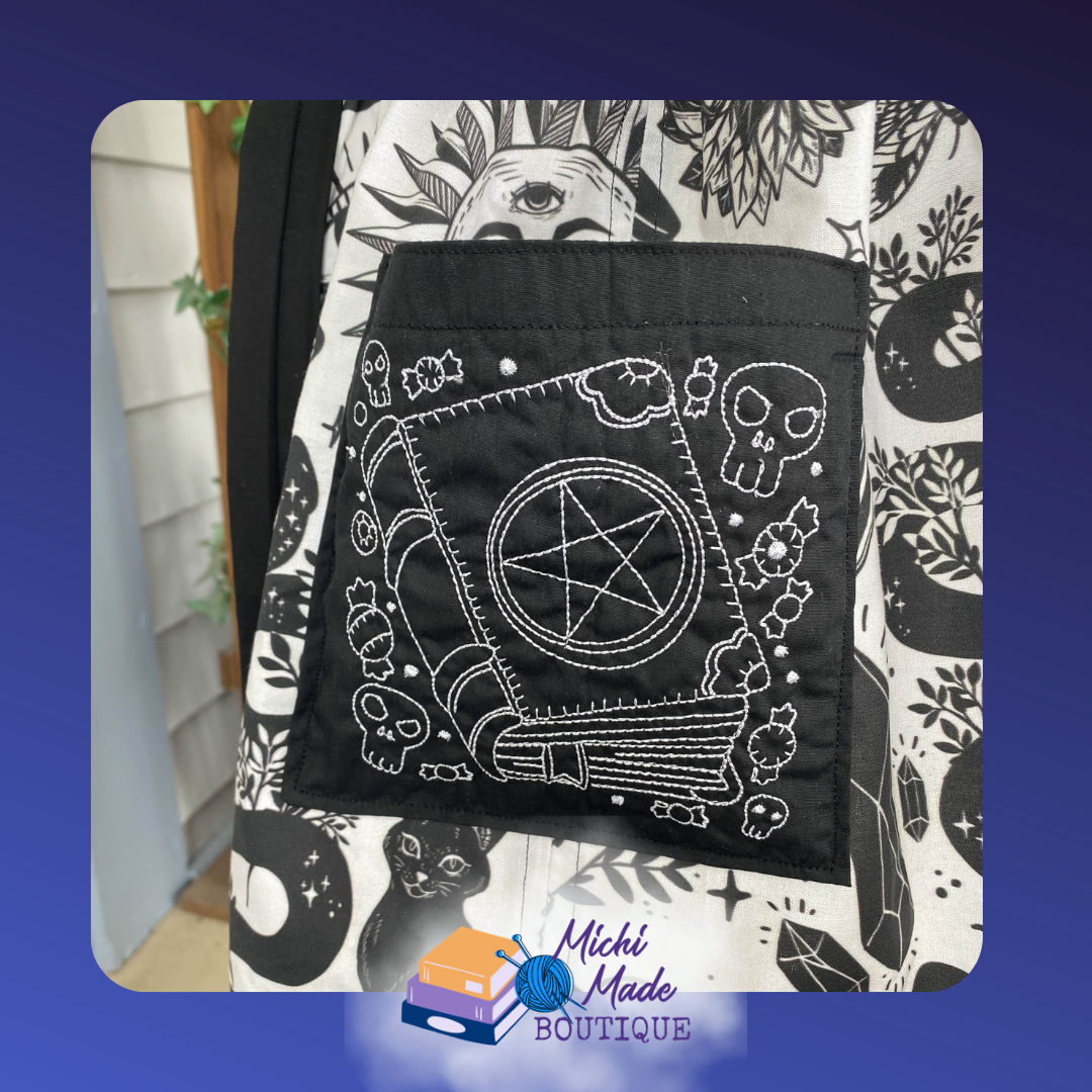 Black apron pocket embroidered in white with a pentacle, crystal wand, skulls, and florals in a witchcraft altar scene.