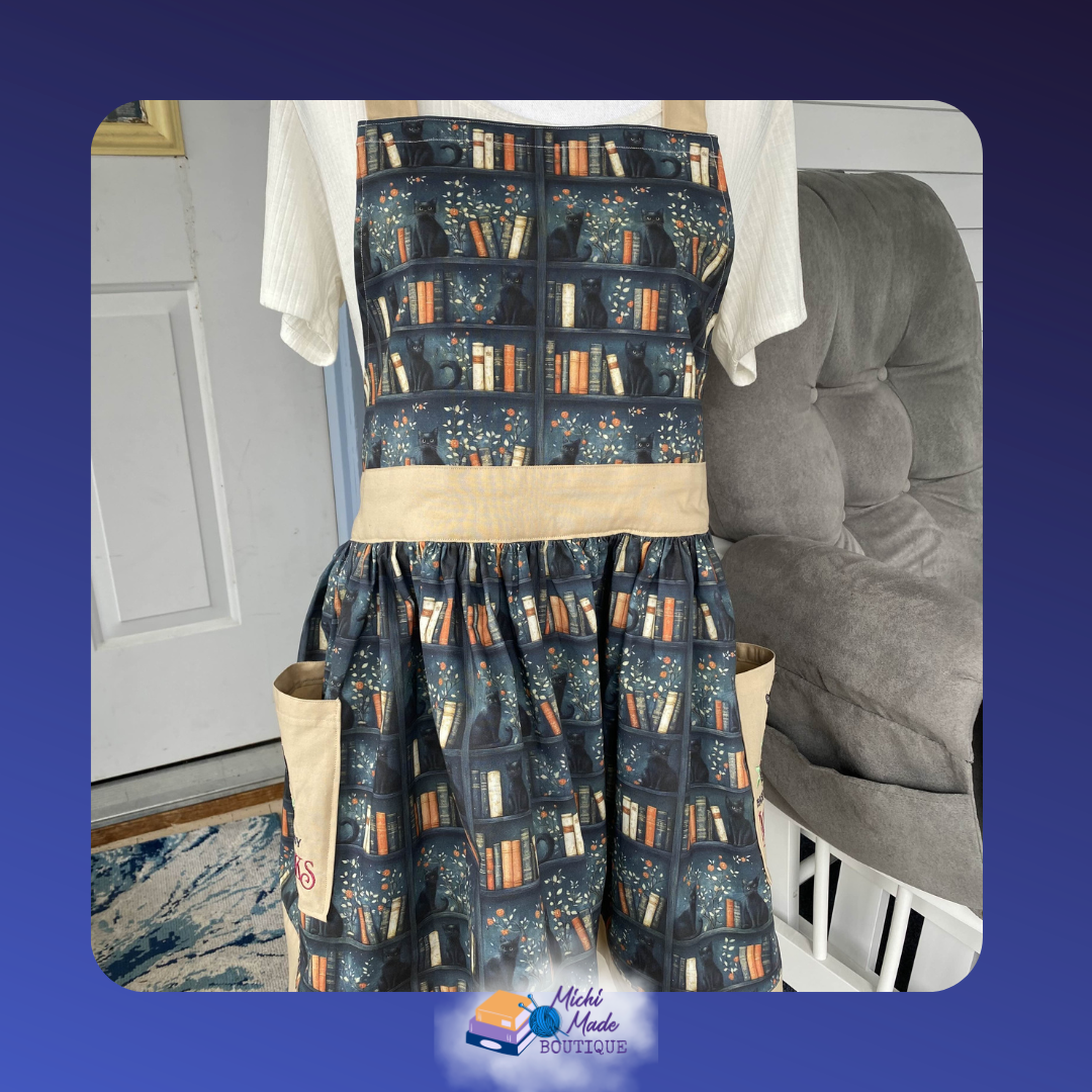 Easily Distracted By Cats and Books Apron.png Dark teal apron with a bookshelf print featuring black cats among colorful books. Cream waistband, gathered skirt, and two cream embroidered pockets.