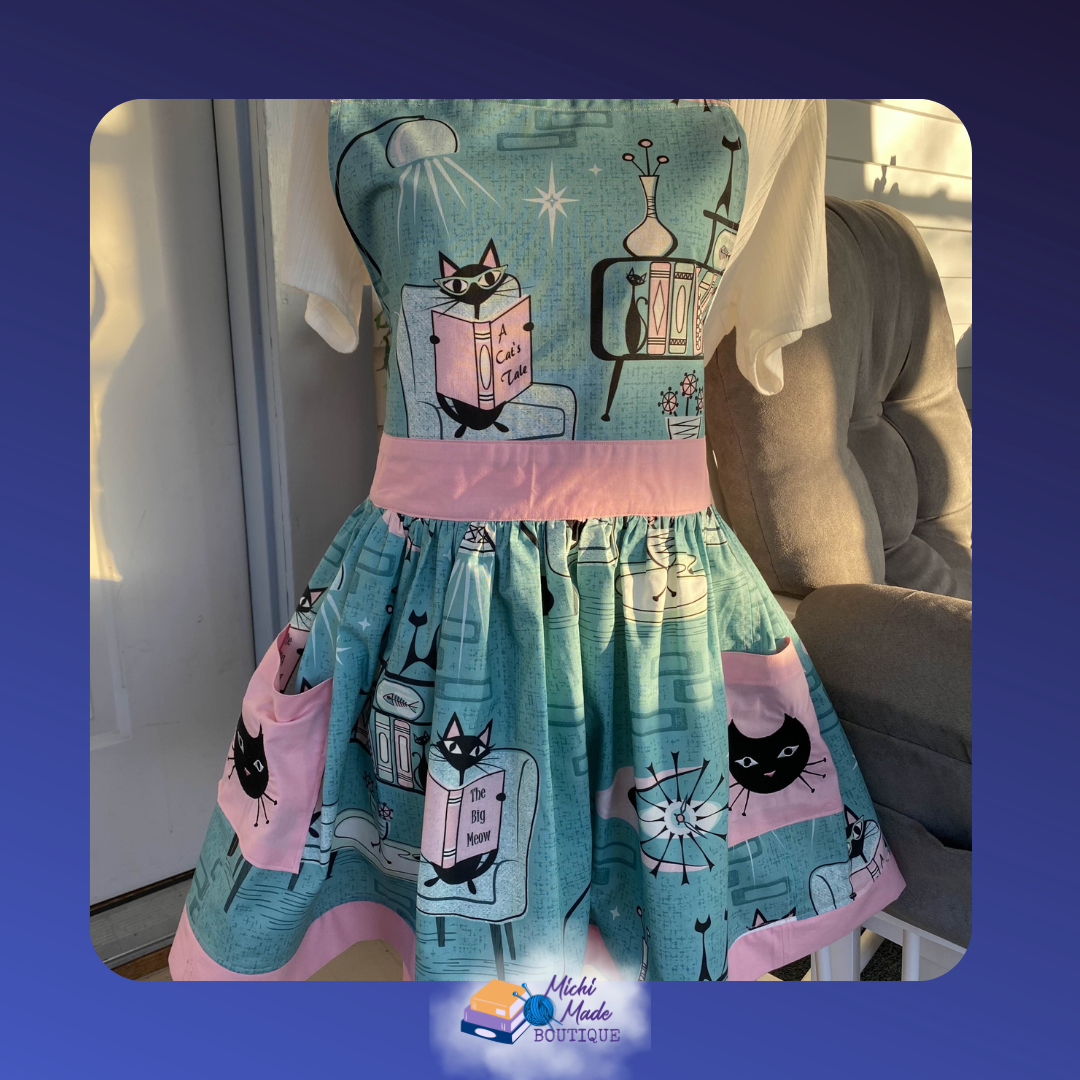 Teal retro apron with mid-century black cats among mod furniture, pink waistband, full gathered skirt, and two pink patch pockets.