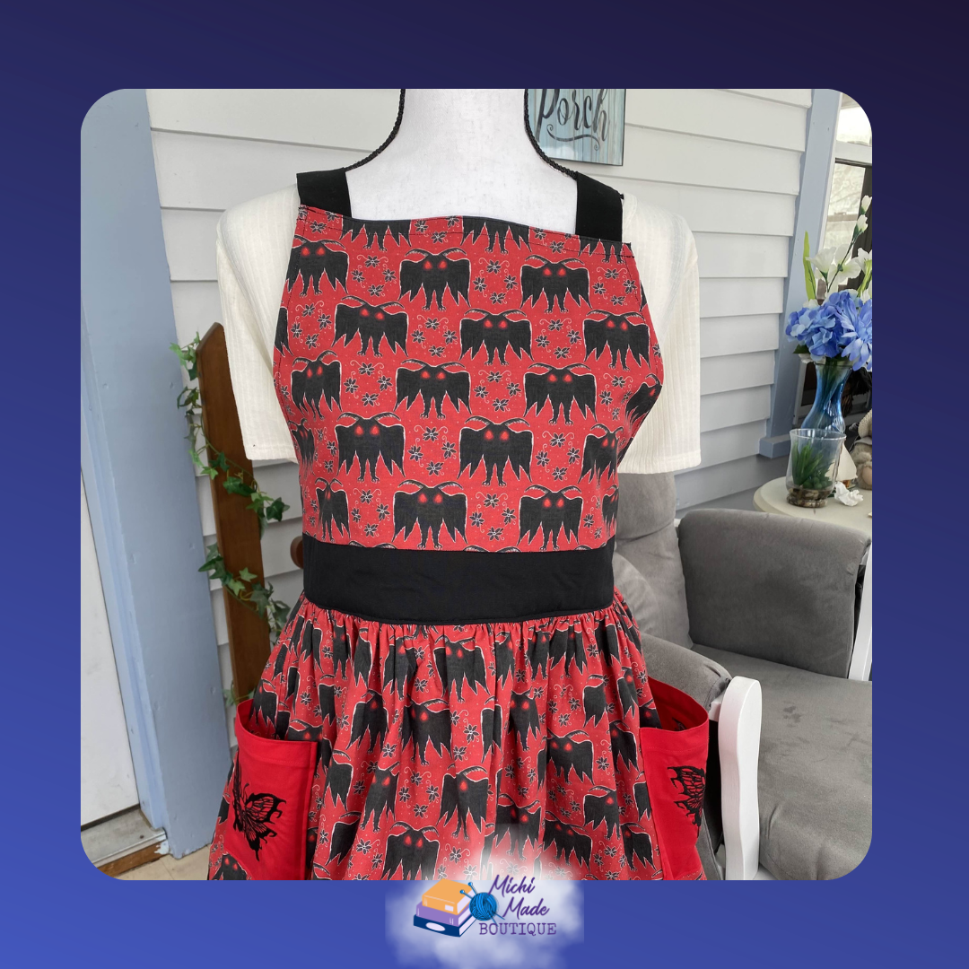 Mothman_1.png Red apron on a dress form covered in a repeating black Mothman silhouette print with glowing red eyes, black straps and waistband, and a full gathered skirt with embroidered red pockets.
