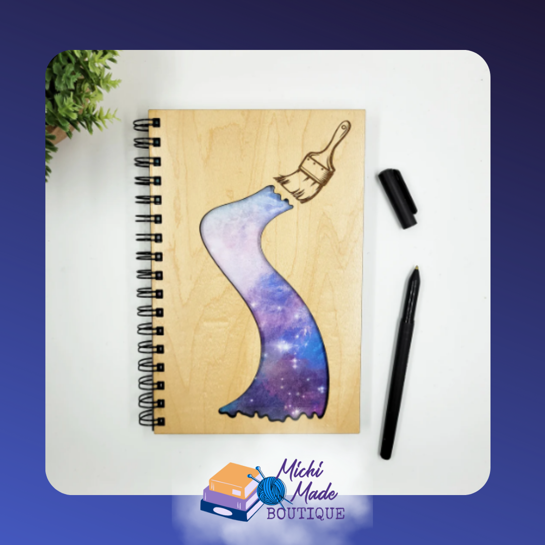 Paint the Sky – Wood journal with engraved paintbrush painting a swirling galaxy of purple, blue, and pink.