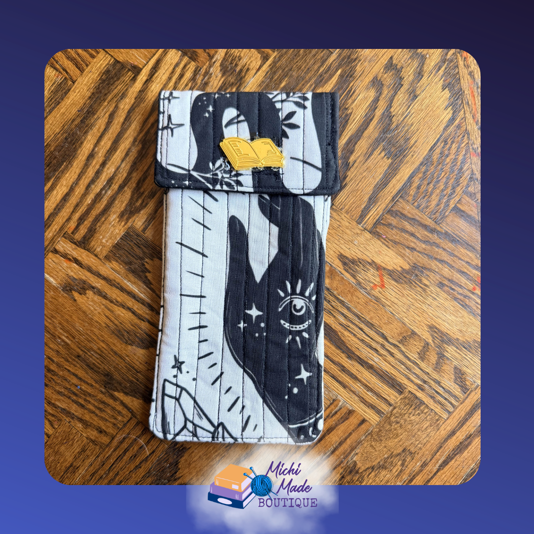 Witchy Eyeglass Case.png Quilted eyeglass case in white and black witchcraft print with a palmistry hand and all-seeing eye, black flap, and yellow open book button.