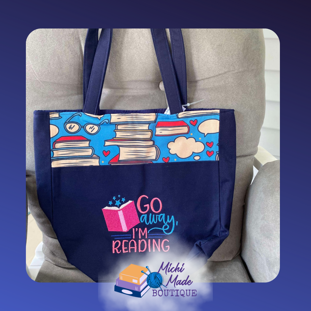 Go Away I'm Reading Embroidered Tote Bag Midnight blue tote bag w/a fabric panel of stacked books and hearts, and Go Away I'm Reading embroidered in pink and teal above a glowing open book.