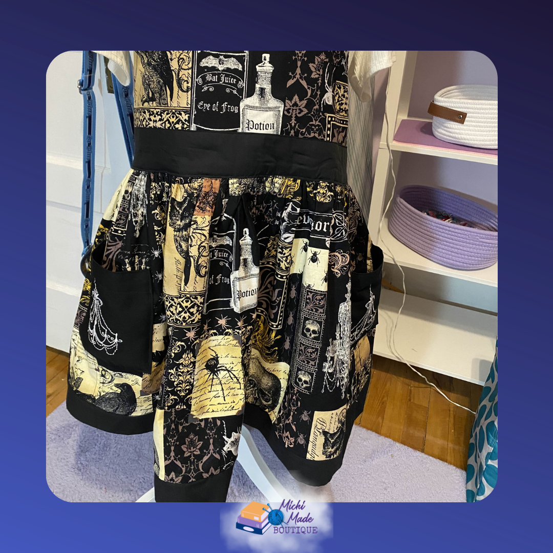 Apothecary_2.png Gathered skirt of the Apothecary Apron with two black patch pockets, showing fabric print details including a spider, black cat, skulls, and damask motifs on cream and black.