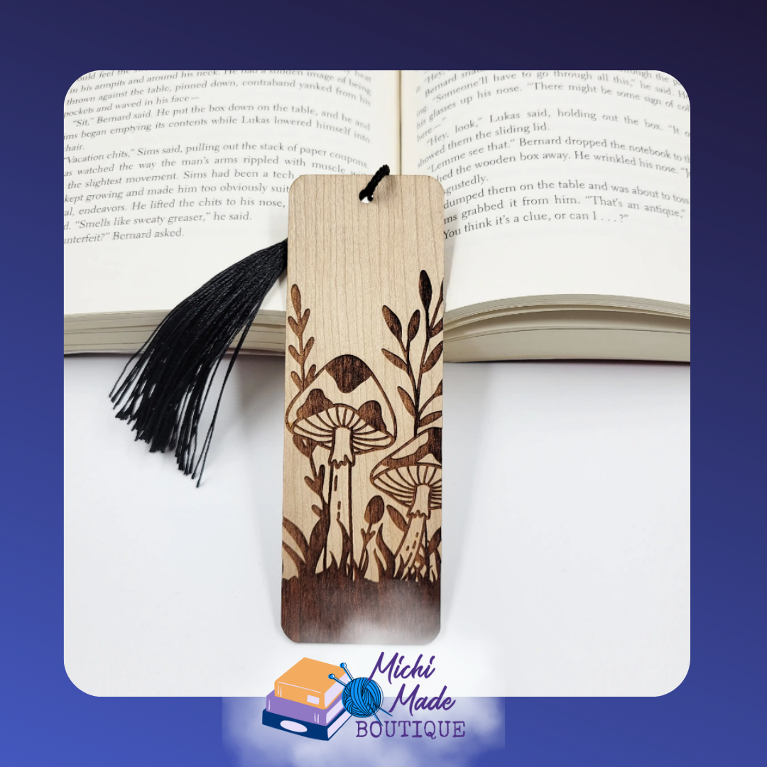 Wooden bookmark with engraved folk art mushrooms and plants, black tassel, on open book.