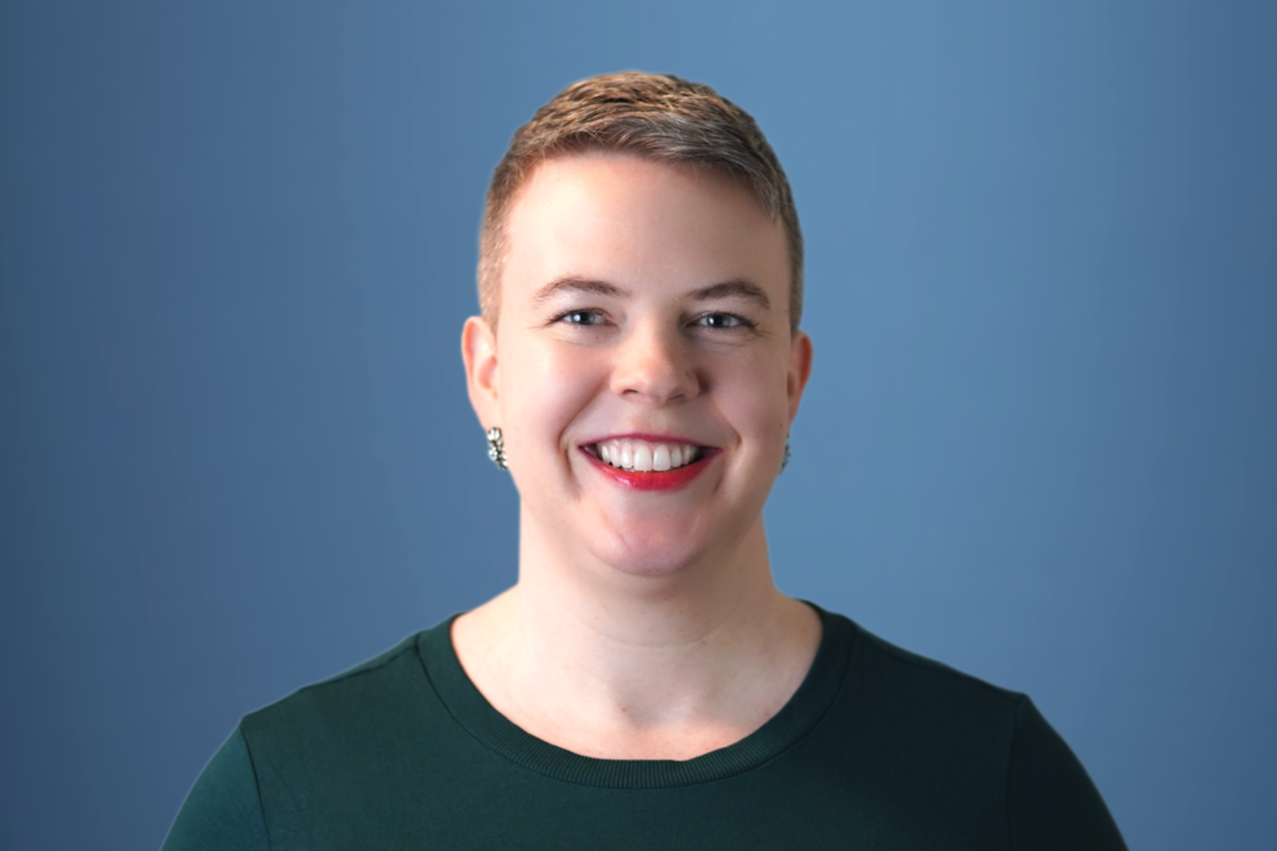 Professional headshot of a woman with dark hair pulled back, wearing a black blazer and light blue shirt, smiling against a dark background.