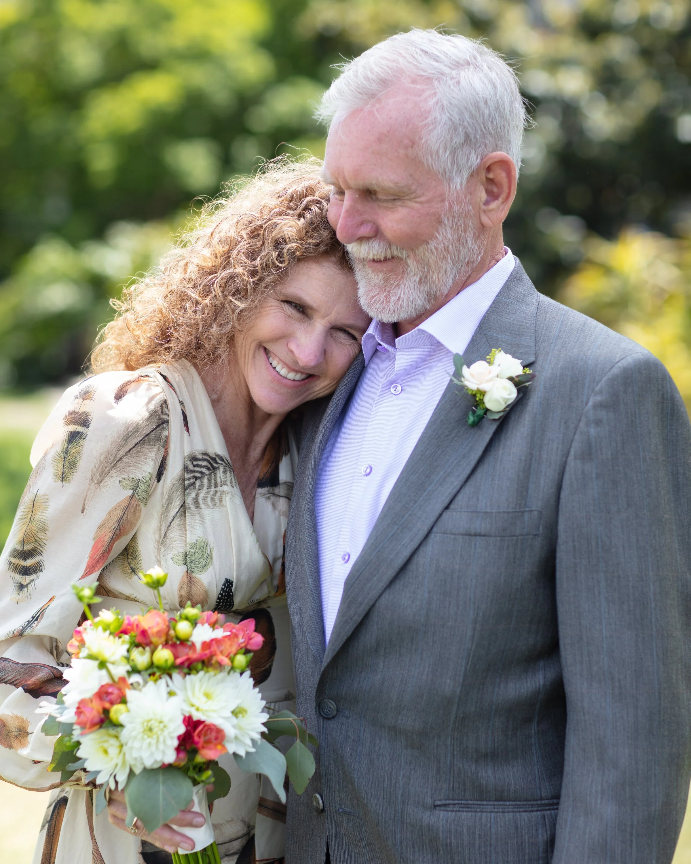 A happy older wedding couple,  outdoors at the Santa Barbara courthouse, smiling at the camera. The 40+ bride holds a colorful bouquet of flowers, and the older groom is wearing a gray suit with a white flower boutonniere.