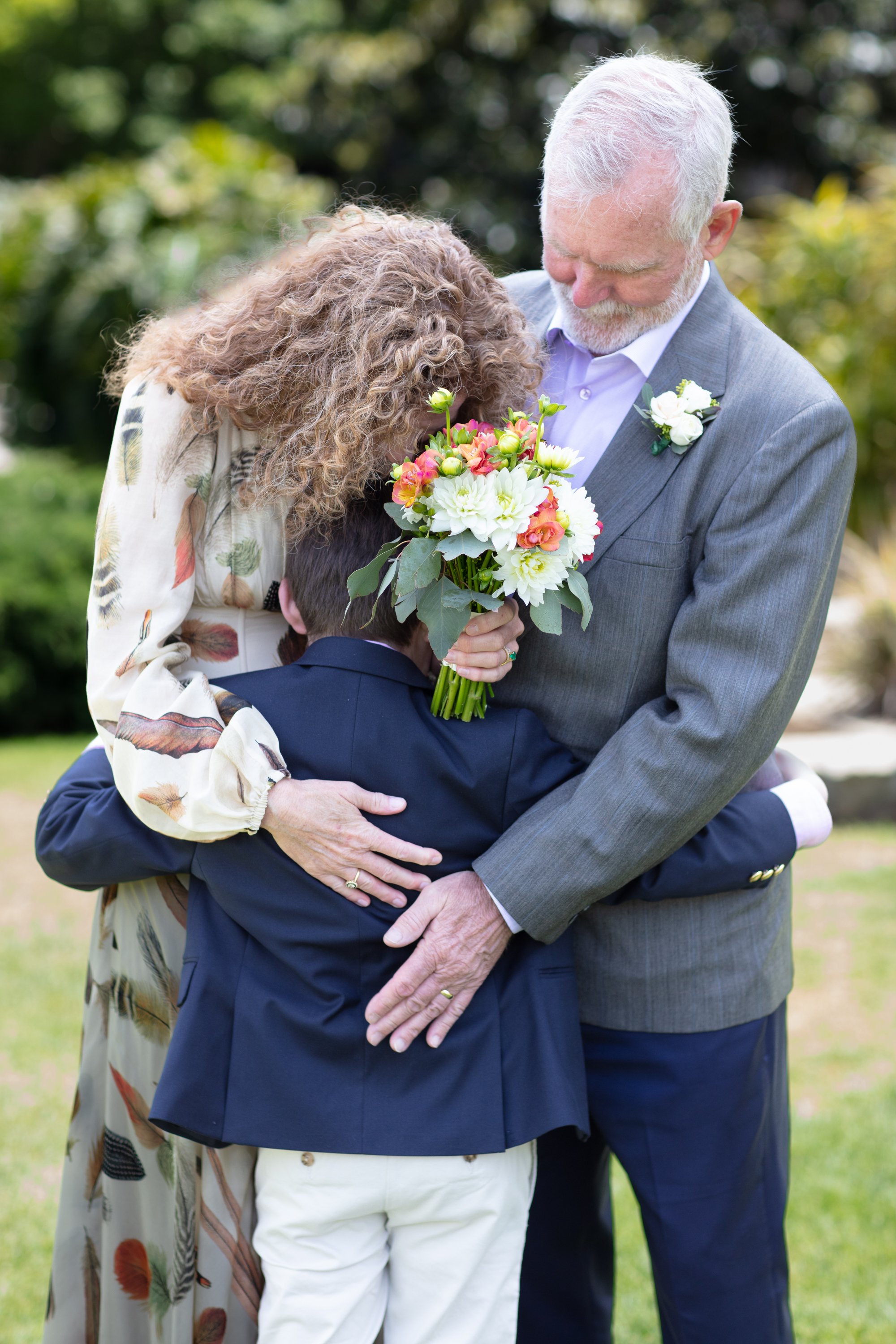 An older groom in a gray suit, an older bride with a floral dress, holding a bouquet of flowers, hug a young boy in a navy blazer outdoors at the Santa Barbara courthouse after their wedding. They are outdoors on a sunny day.