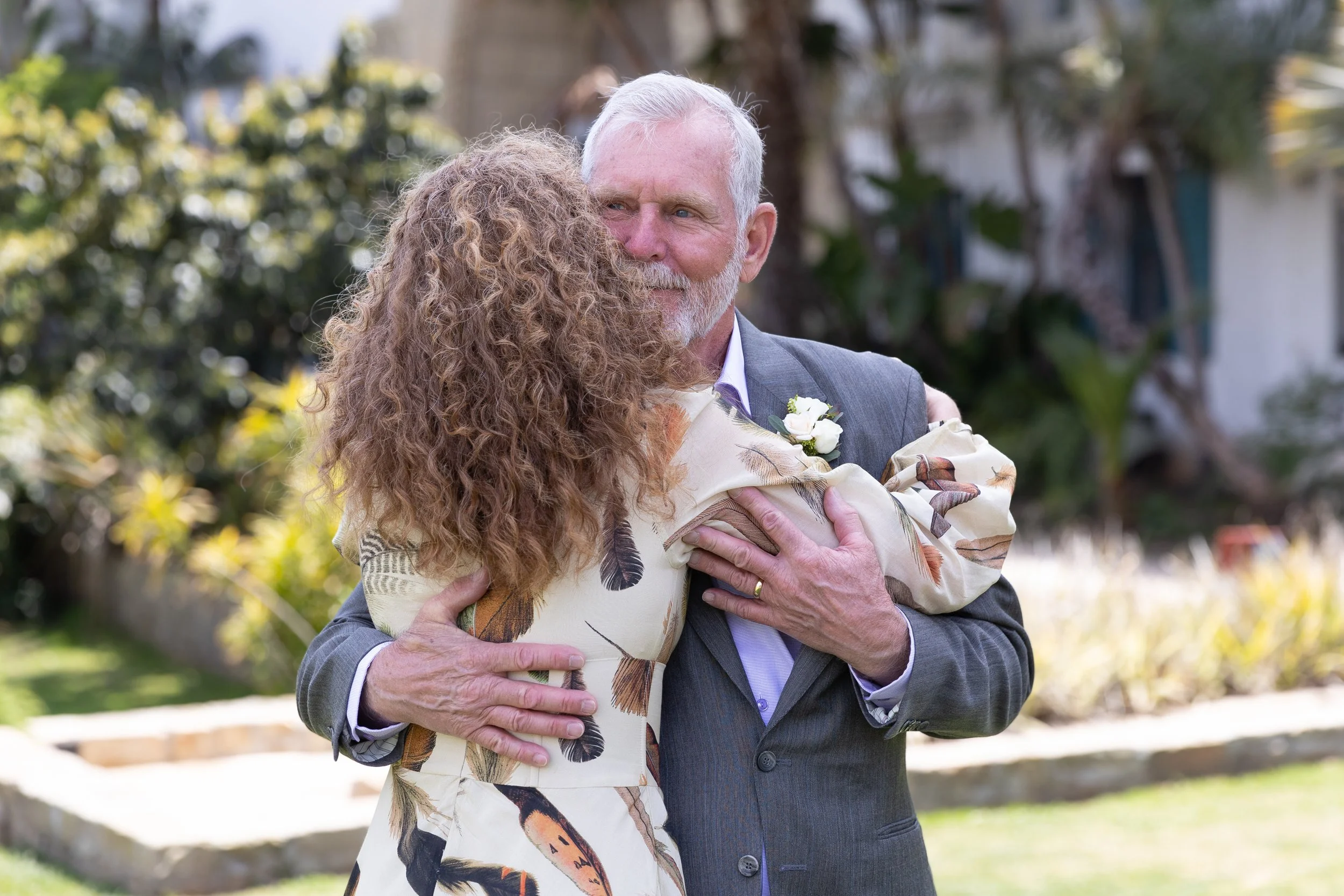 An older 40+ couple after their wedding ceremony at the Santa Barbara courthouse  hugging outdoors, with the man with white hair and beard, smiling and wearing a gray suit, and the woman in a dress with a feather pattern.