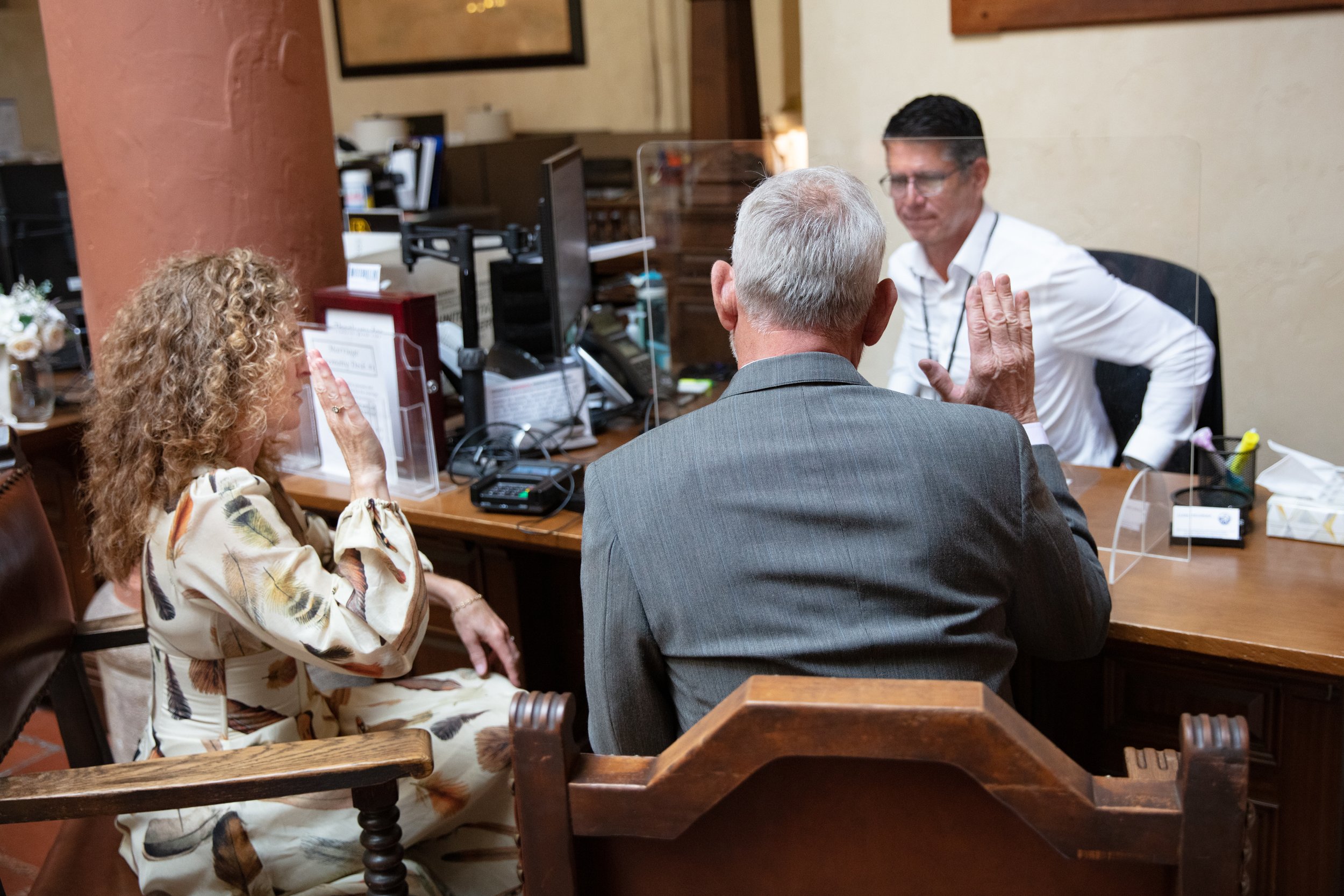 A woman and a man are sitting at a desk with a wedding officiant, saying their wedding vows. The woman appears emotional with her hand raised, groom is raising his hand also.