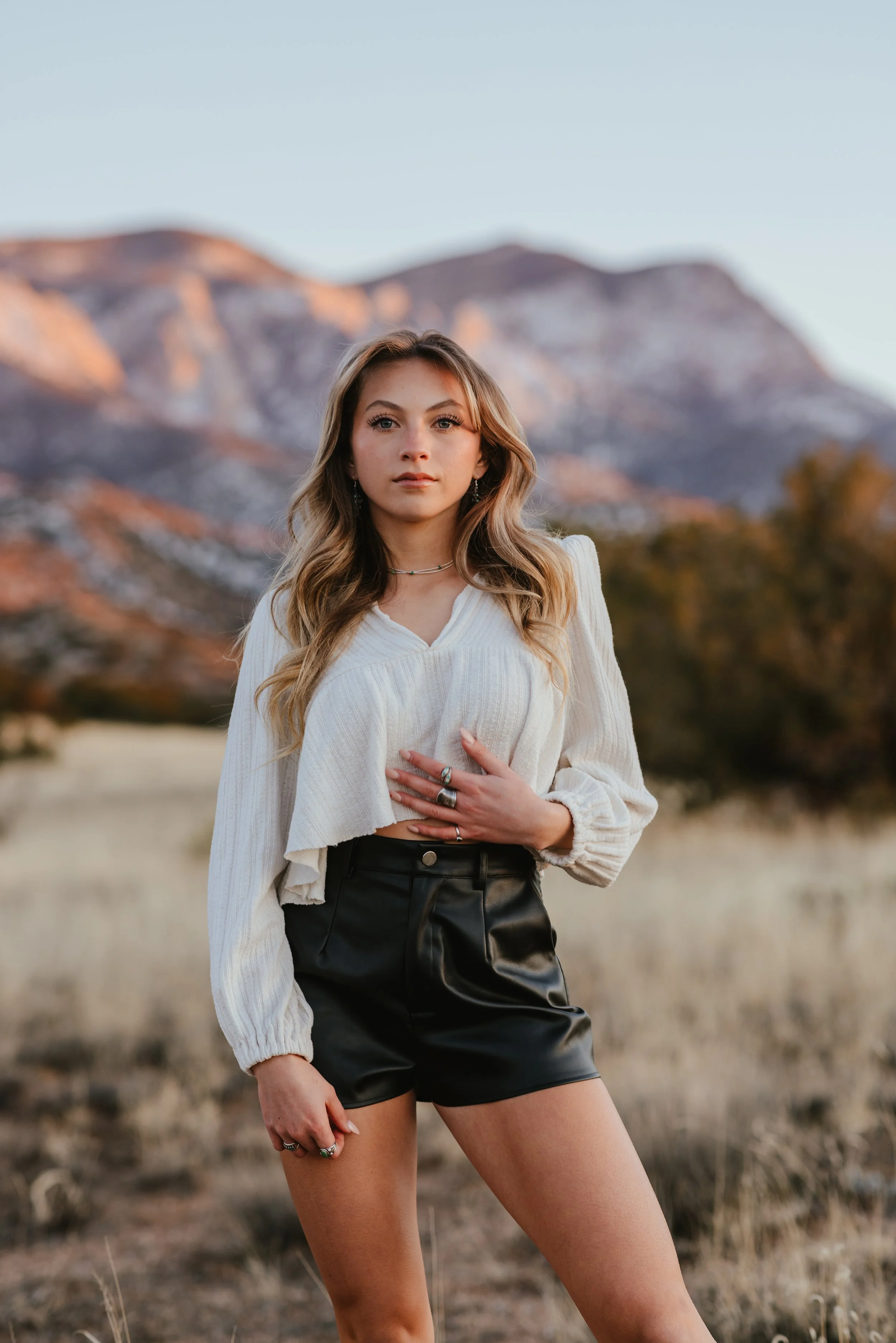 A woman in a white long-sleeve top and black shorts stands in a grassy field with mountains in the background.
