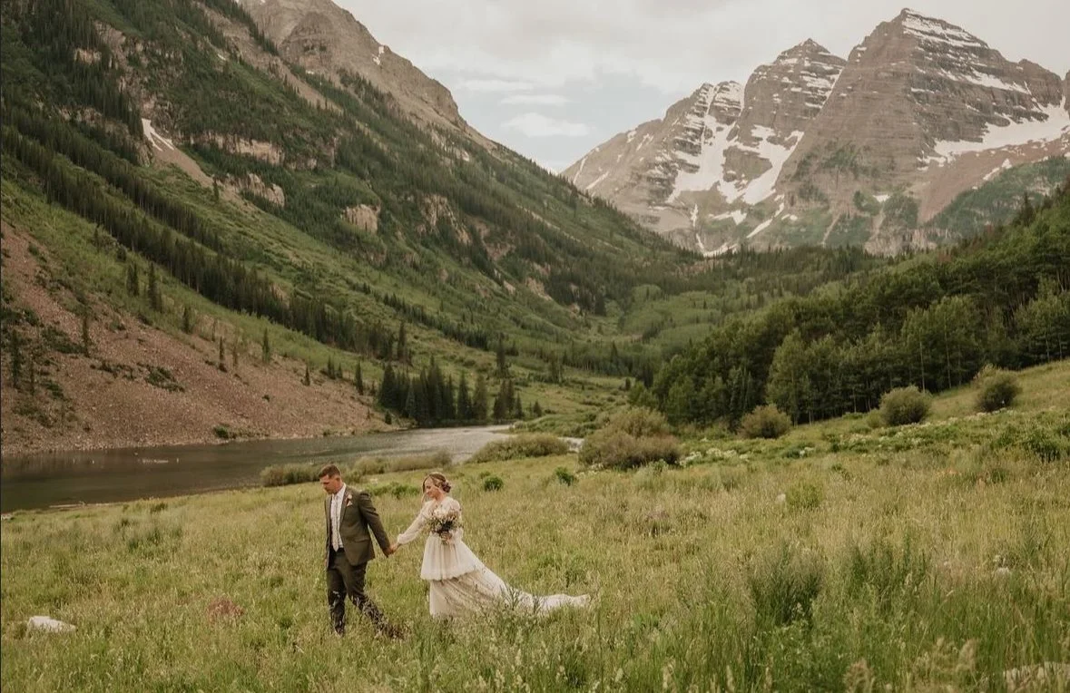 Bride and groom walking hand in hand through a grassy field with mountain and lake backdrop.