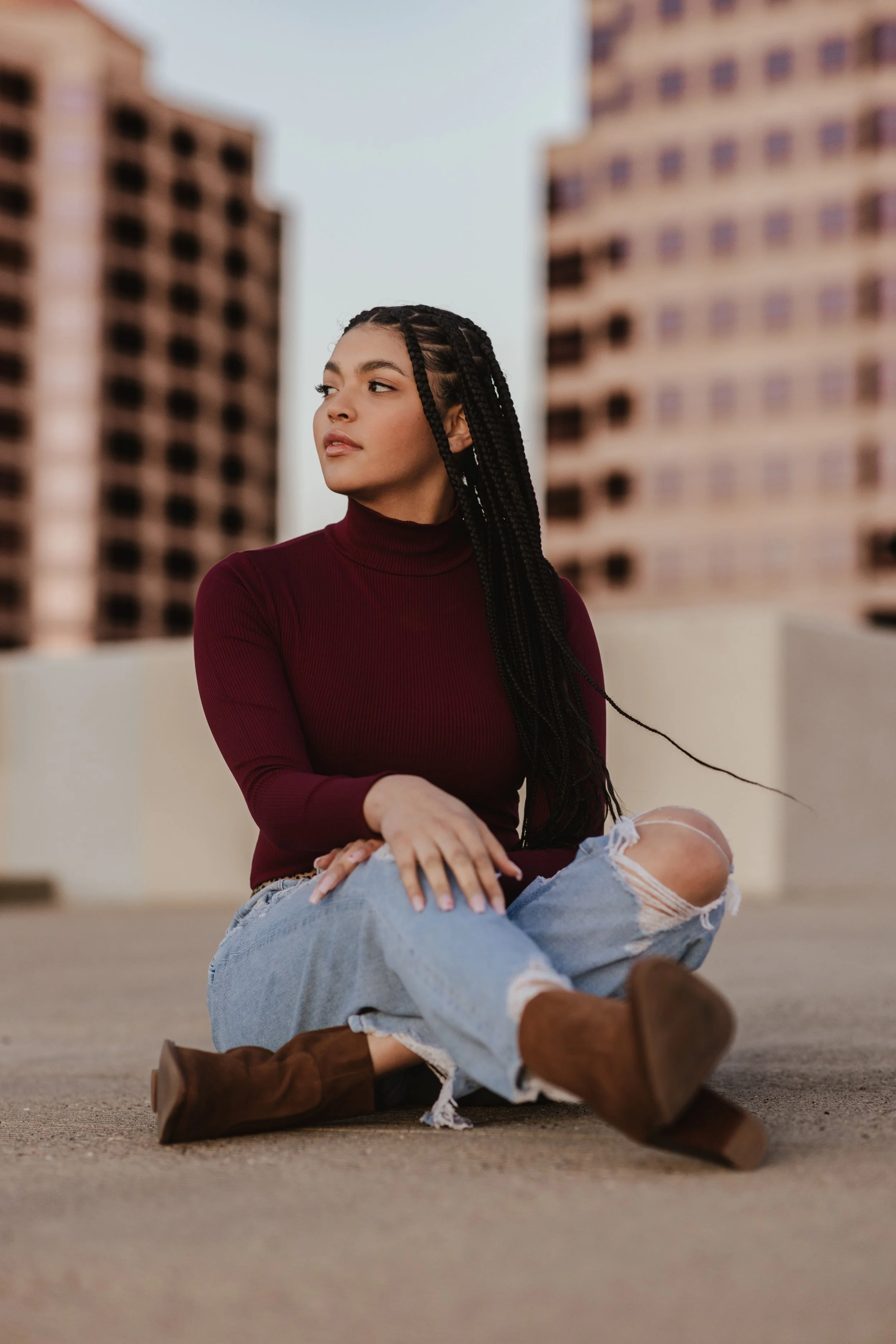 Woman with long braided hair sitting on ground, wearing maroon turtleneck, ripped jeans, and brown boots, with blurred buildings in the background.