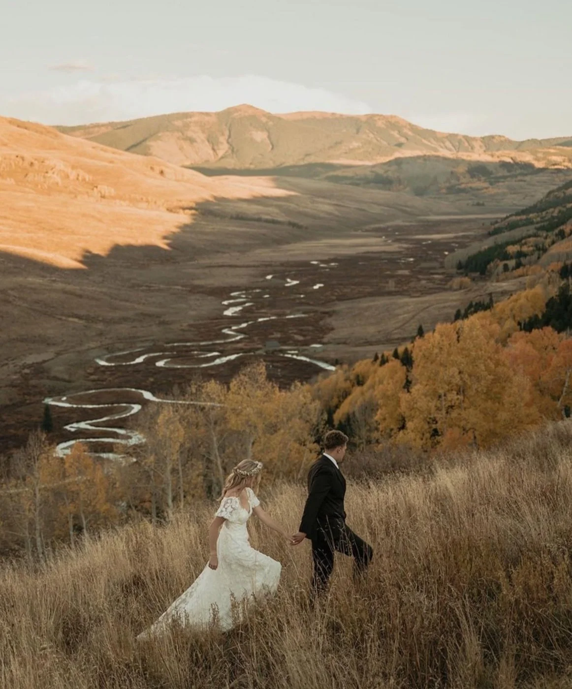Bride and groom walking through a grassy hill with a winding river and mountain landscape in the background, with autumn foliage.