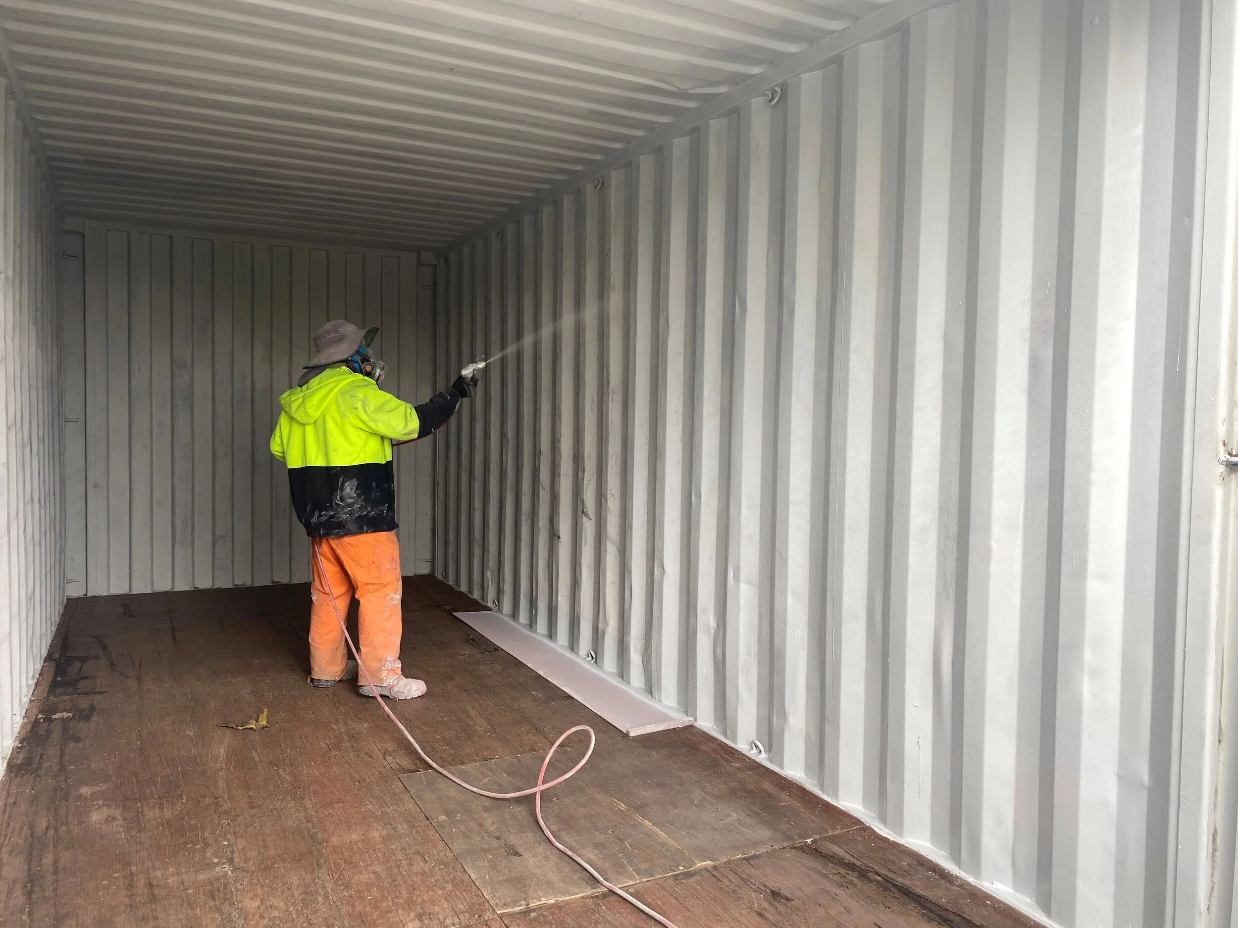 A person wearing a neon yellow and black jacket, orange pants, and a wide-brimmed hat is spray painting the interior of a shipping container.
