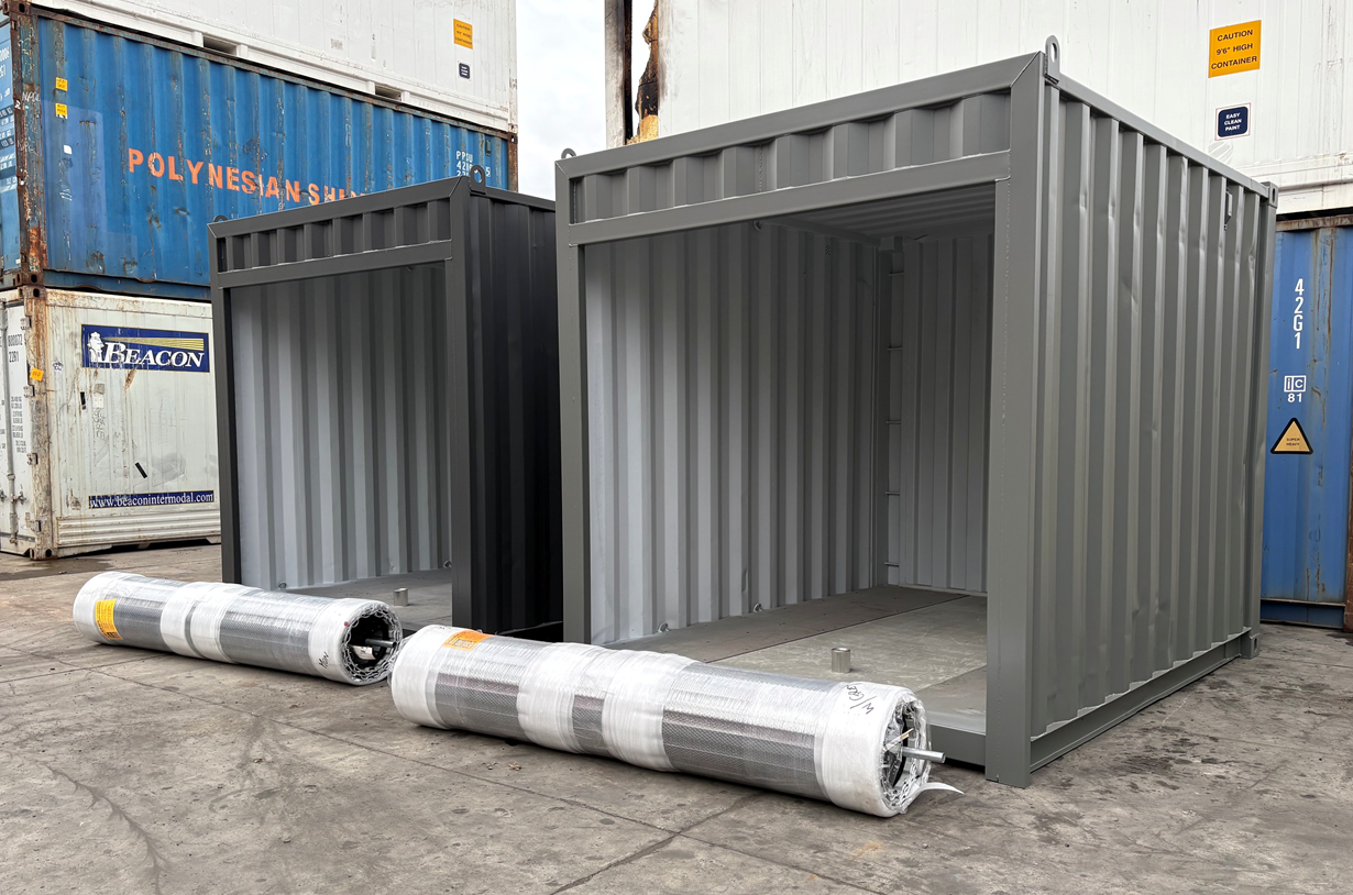 Two 10ft shipping containers, one dark grey with open side, and one light grey with closed side waiting for roller doors to be fitted