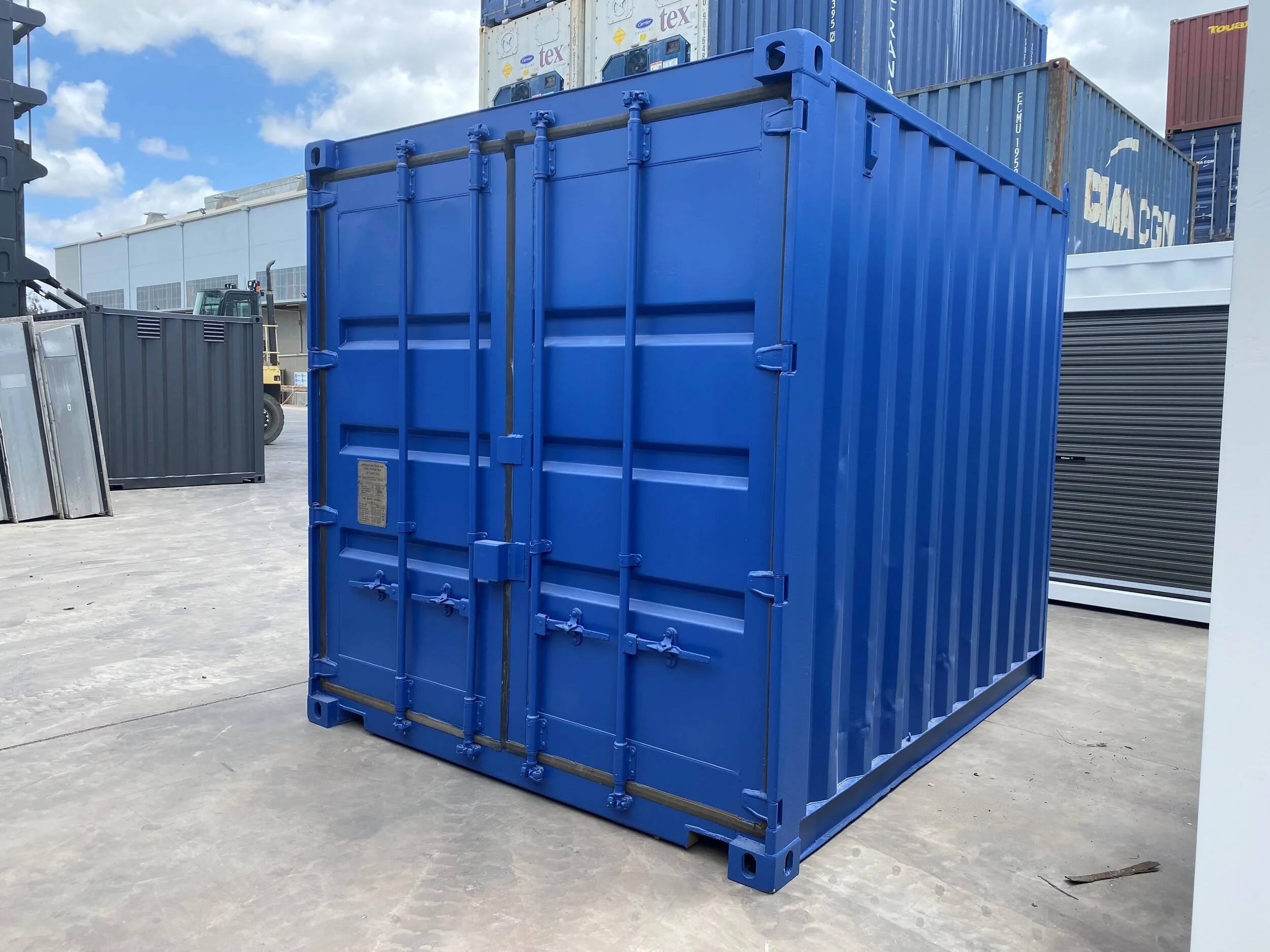 A large blue shipping container on concrete surface, with other containers and a forklift in the background, under a partly cloudy sky.