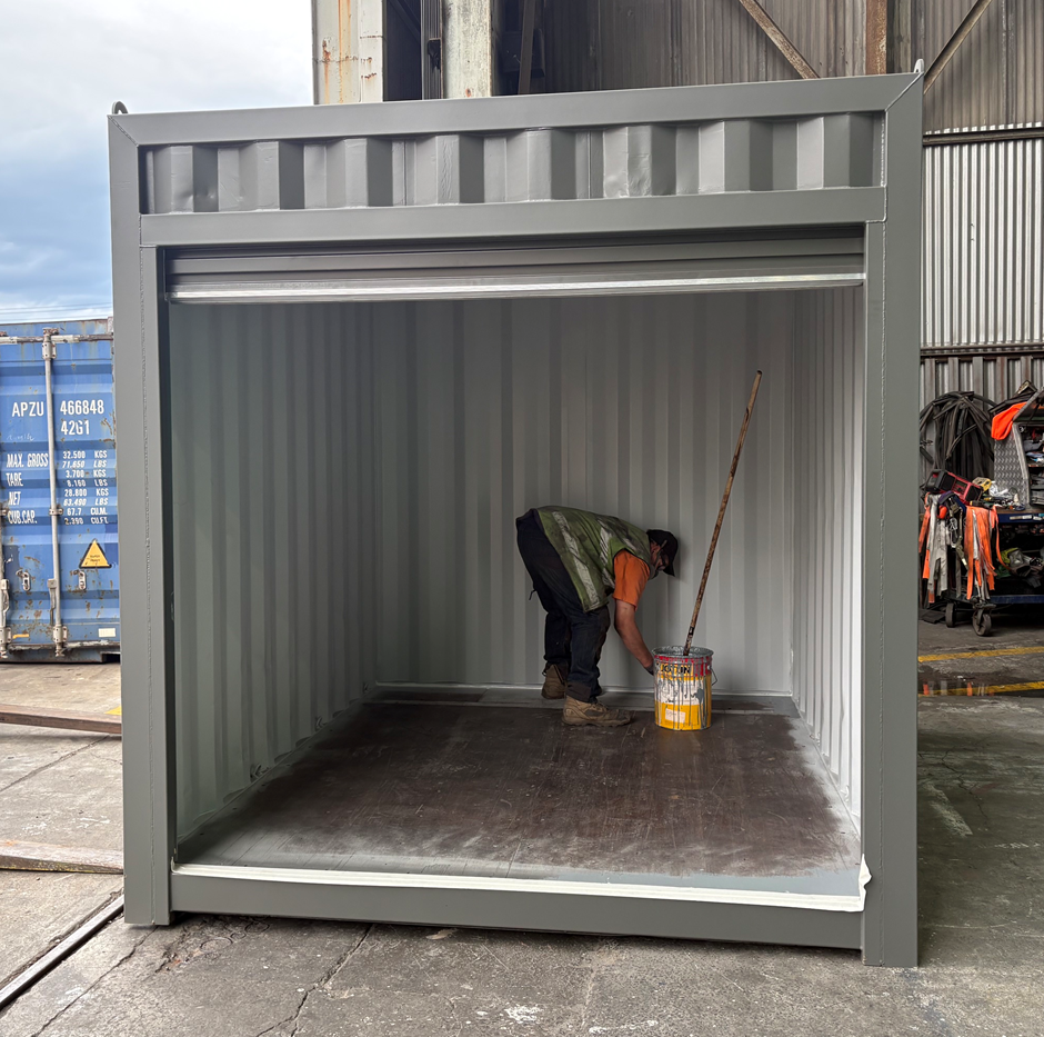 A man in an orange shirt and black vest is painting the interior of a gray shipping container with a roller brush, standing on a wooden floor and holding a paint bucket inside a 10ft shipping container