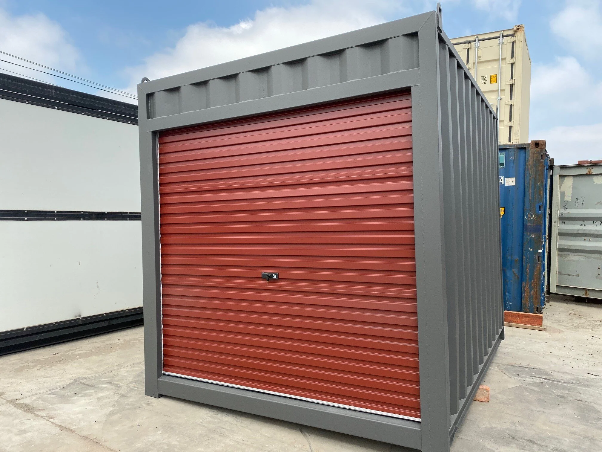 A 10ft grey container with a red roller door, situated outdoors on a concrete surface, with other similar containers in the background and a partly cloudy sky overhead.