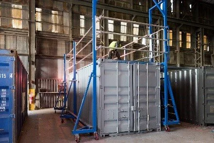 Shipping container workshop worker standing on a platform scaffolding next to a large gray metal container inside a warehouse or industrial building.