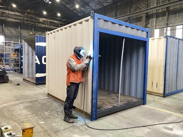 Worker welding or assembling a shipping container inside a warehouse, wearing protective gear including a helmet and safety vest.