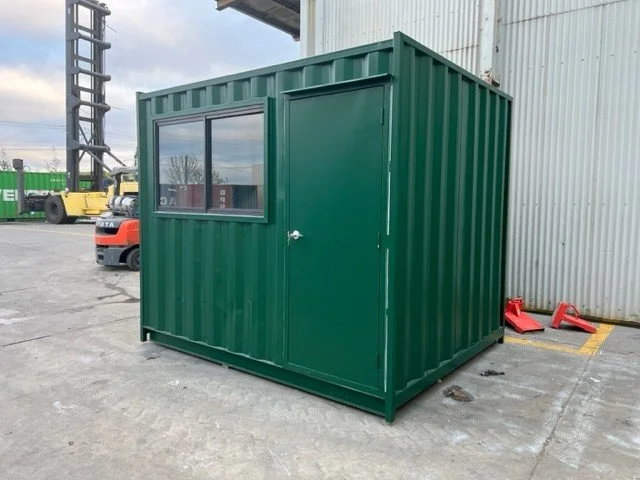 A custom sized green shipping container shed with a window and a door, placed outdoors near a warehouse.