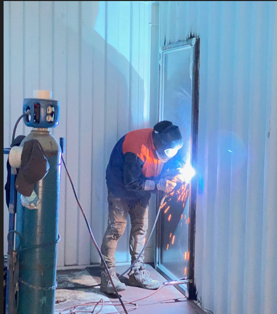 A worker in safety gear welding a metal door frame inside a shipping container