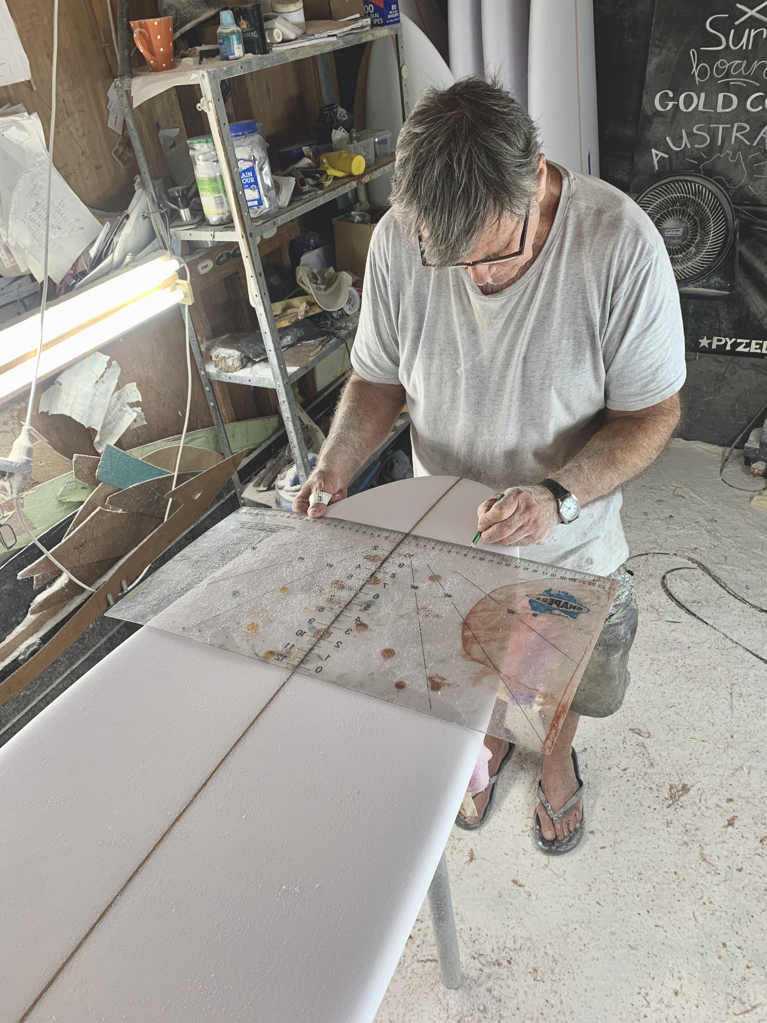 Man shaping a surfboard in a workshop with tools and materials.