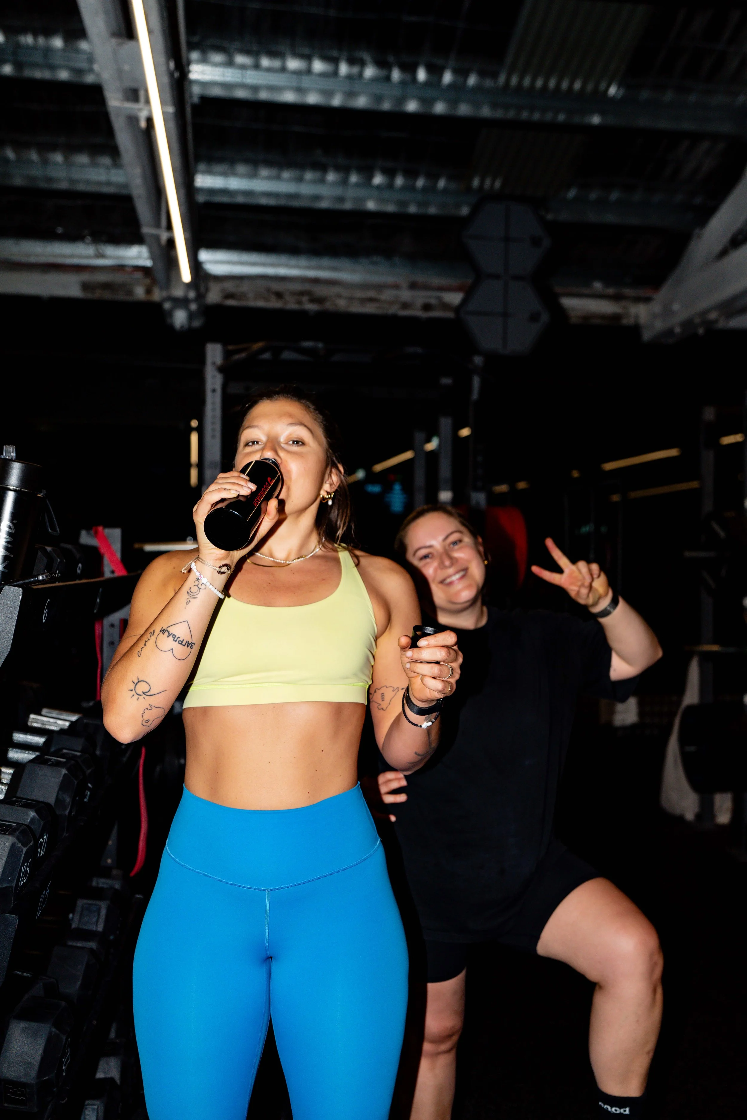 Two women at a gym, one in workout attire drinking from a water bottle, the other smiling and making a peace sign behind her.