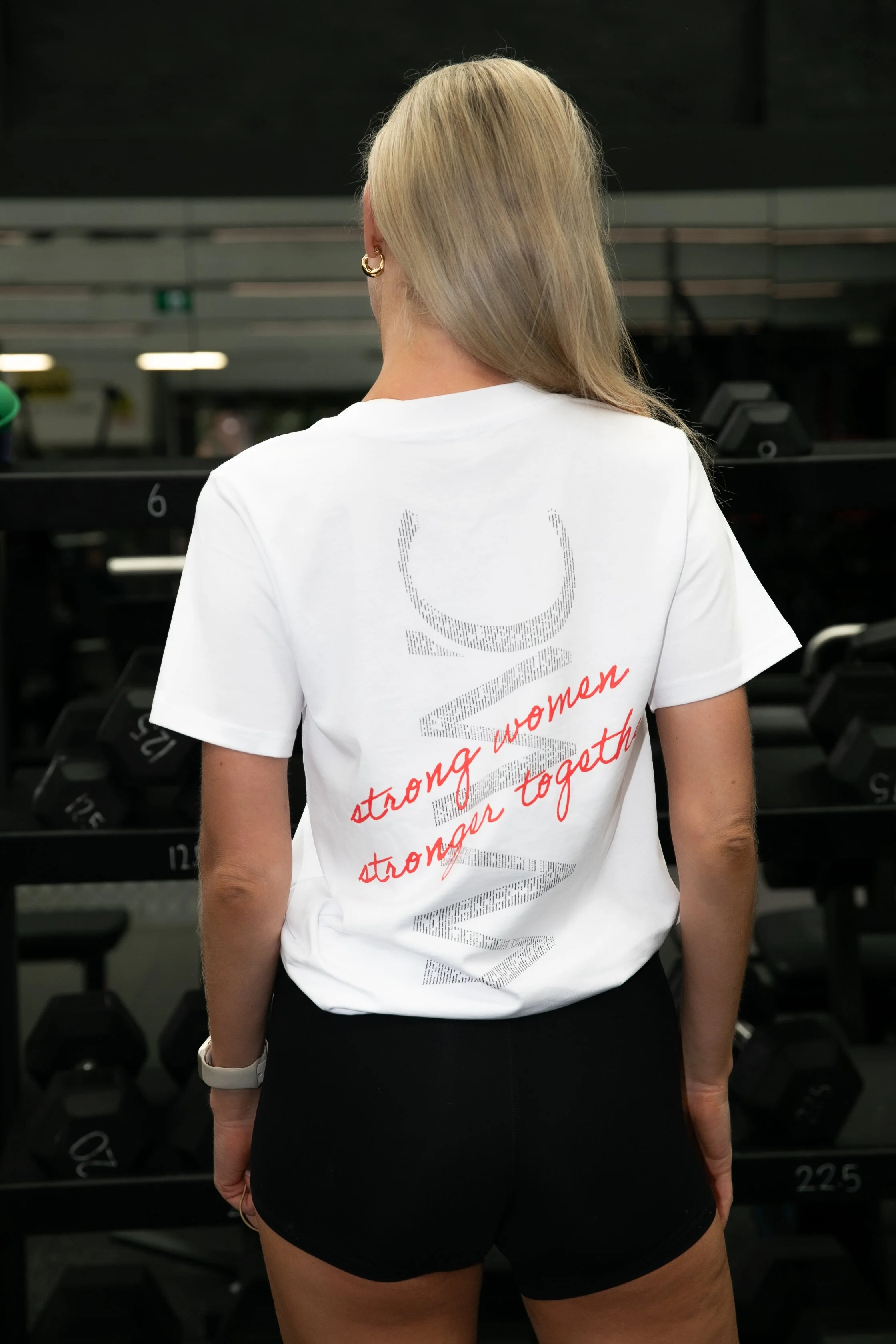 Back view of a woman with blonde hair wearing a white t-shirt with red text and black shorts, standing in a gym with weights in the background.