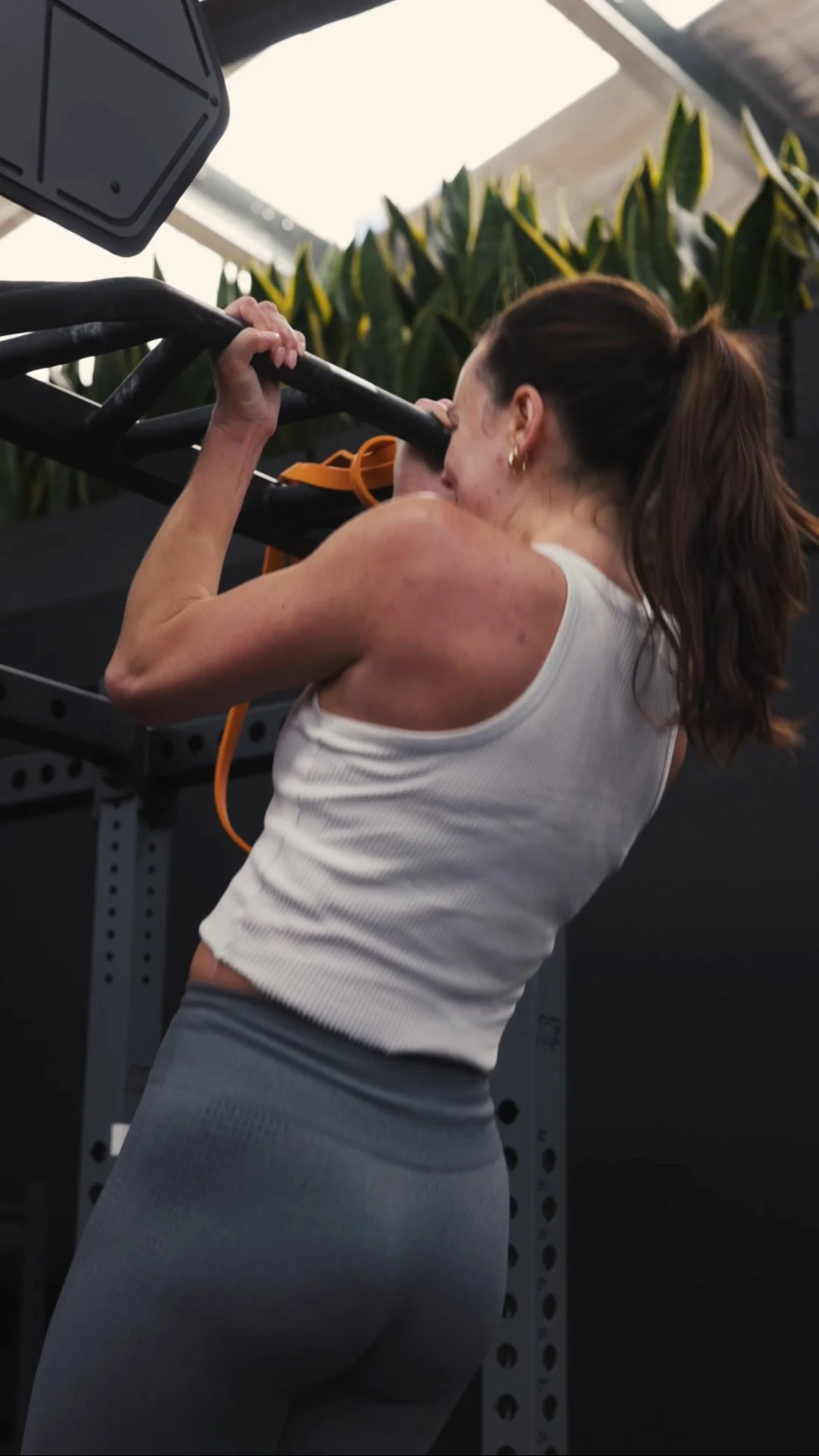 woman in white tank top and gray workout leggings doing pull-ups in gym