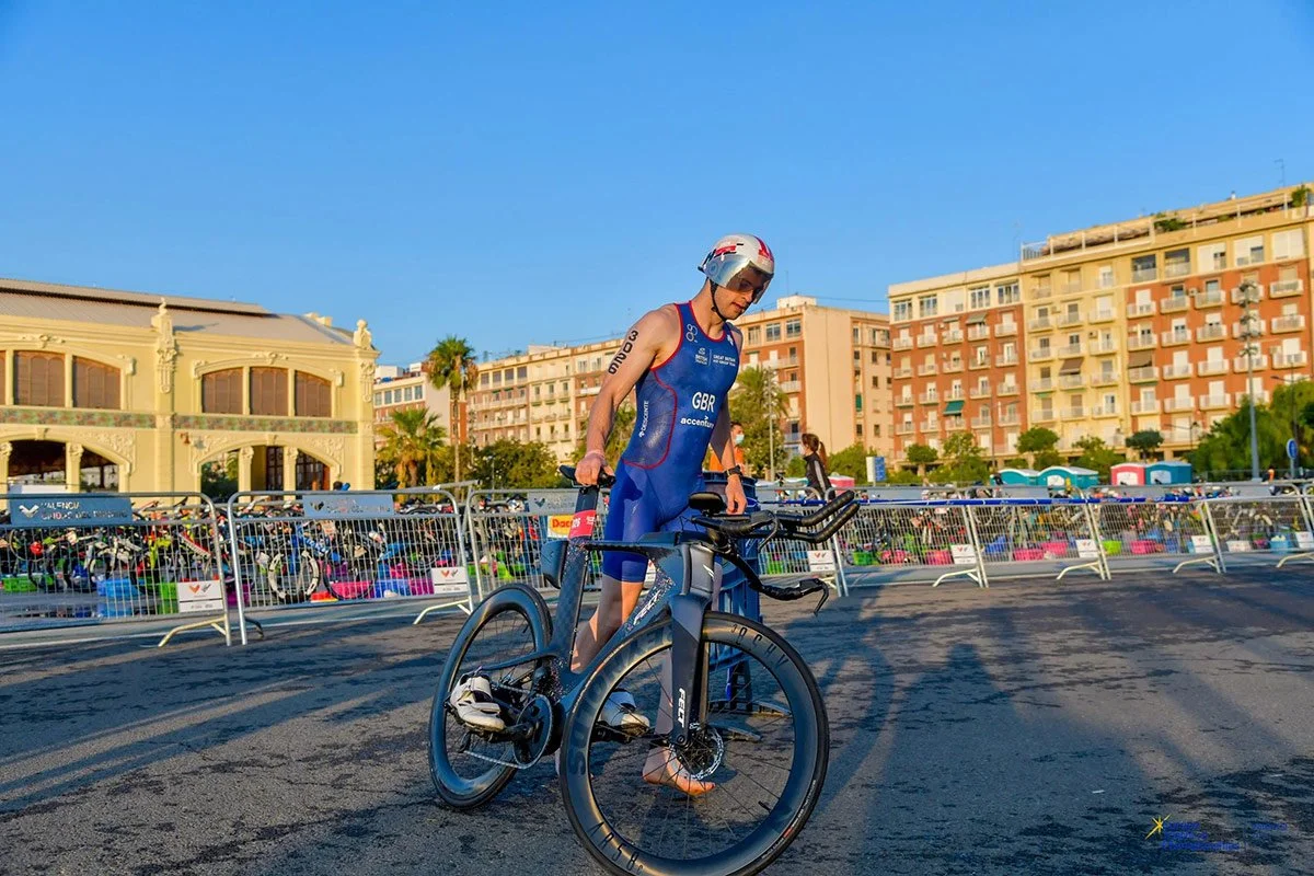 A triathlete in a race suit walks a time trial bike through a fenced transition area at an outdoor event.