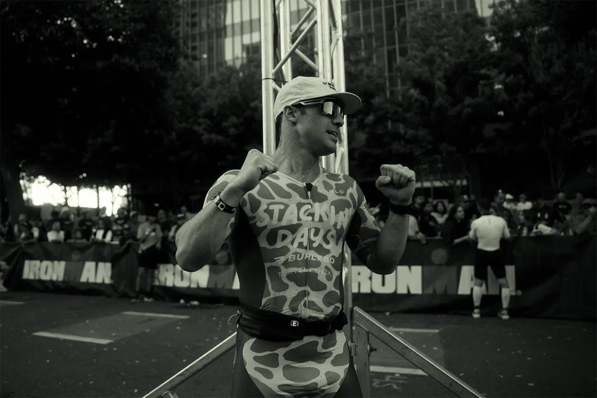 A triathlete in a patterned racing suit stands near the finish area, raising both fists in celebration with a crowd and city buildings behind him.