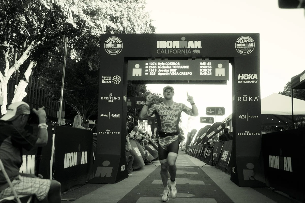 A triathlete runs under the Ironman California finish arch, lifting both arms as spectators line the course.