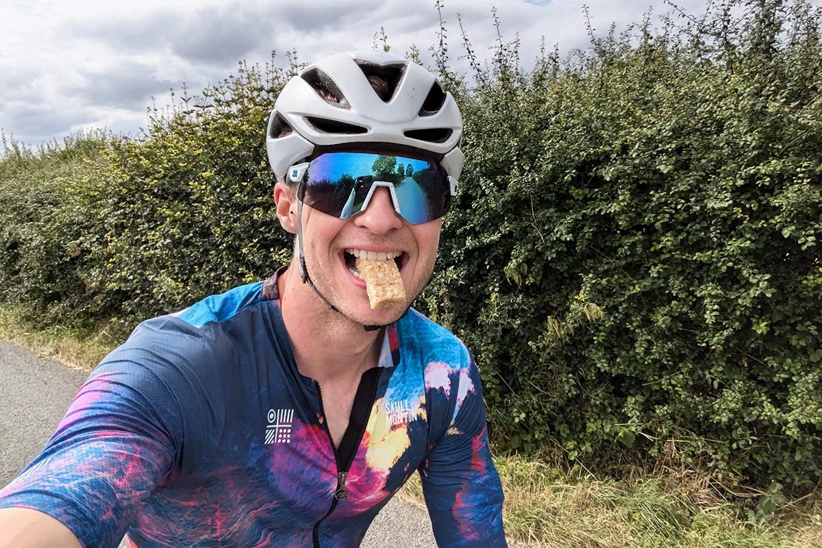 Cyclist wearing a helmet and sunglasses riding outdoors while eating an energy bar.