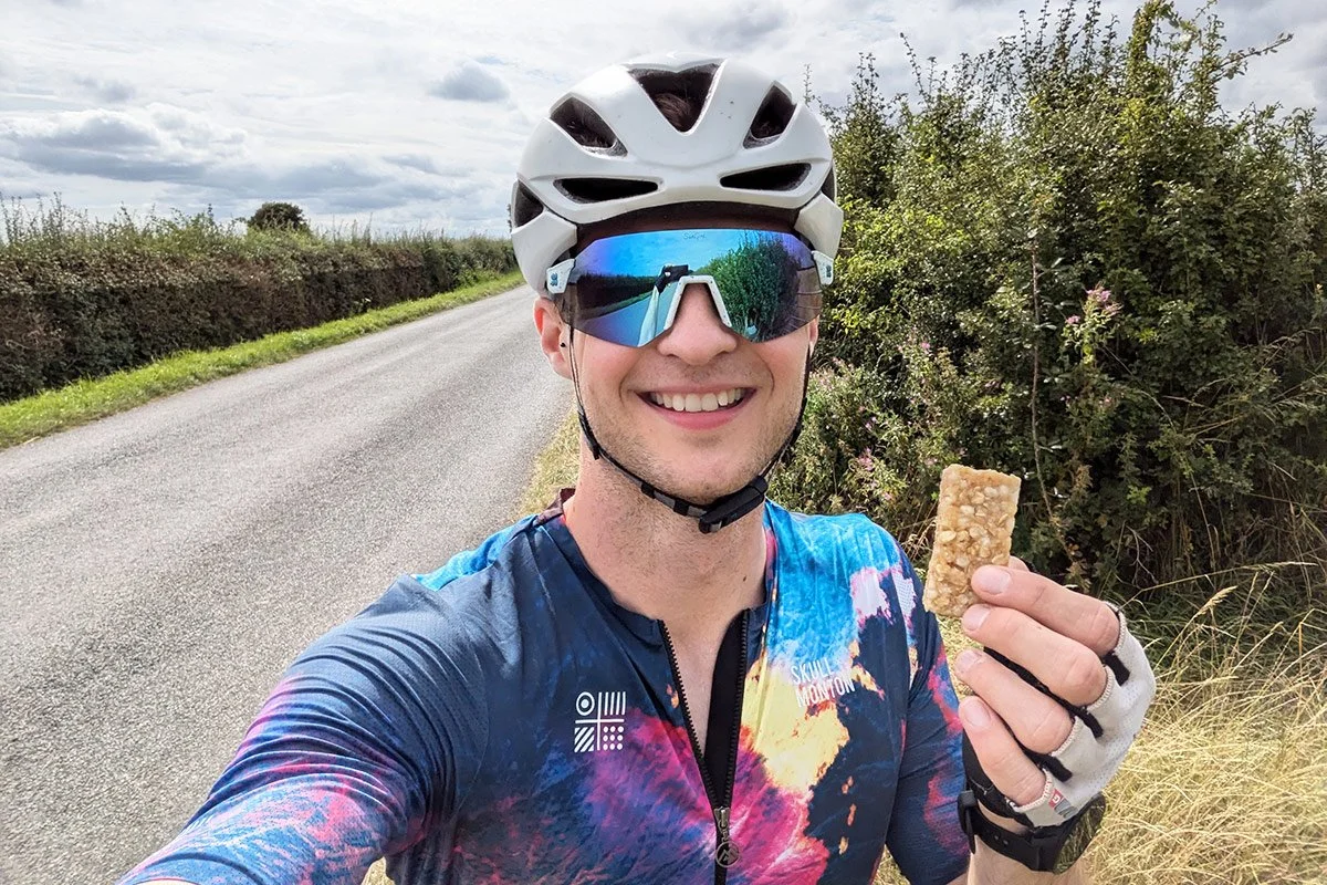 A smiling cyclist wearing a helmet and reflective sunglasses takes a selfie while holding an energy bar on a quiet country road.
