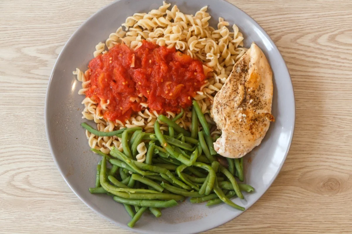 A plate with grilled chicken breast, pasta topped with tomato sauce, and a side of green beans on a light wooden table.