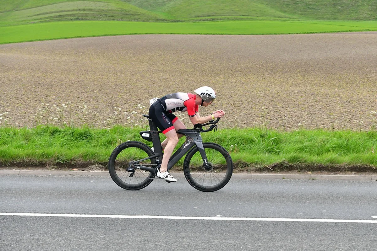 A cyclist in a red and black kit rides a time trial bike on a rural road beside green fields.